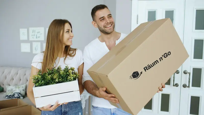 Paar trägt einen Umzugskarton und eine Pflanzenkiste in eine neue Wohnung